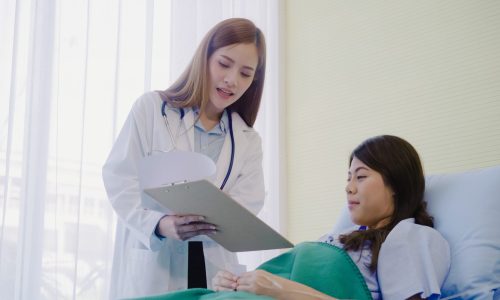 Beautiful smart Asian doctor and patient discussing and explaining something with clipboard in doctor hands while staying on Patient's bed at hospital. Medicine and health care concept.