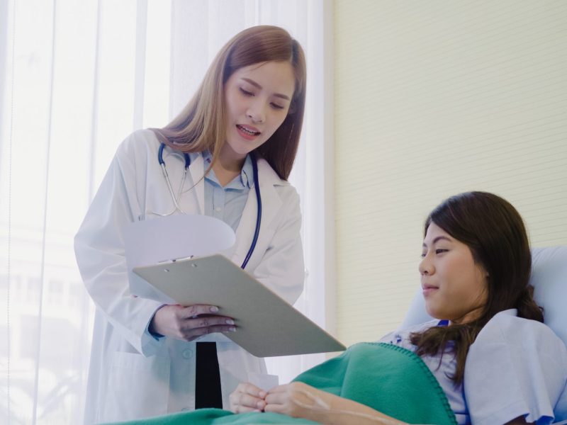 Beautiful smart Asian doctor and patient discussing and explaining something with clipboard in doctor hands while staying on Patient's bed at hospital. Medicine and health care concept.