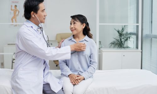 Profile view of confident middle-aged therapist wearing white coat using stethoscope while examining senior patient, interior of modern ward on background