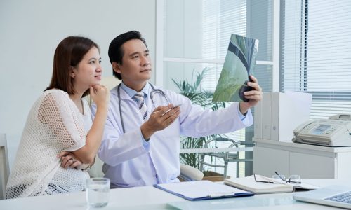 Confident middle-aged surgeon sitting at desk and explaining X-ray results to pretty Asian patient, interior o modern office on background