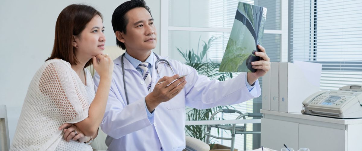 Confident middle-aged surgeon sitting at desk and explaining X-ray results to pretty Asian patient, interior o modern office on background